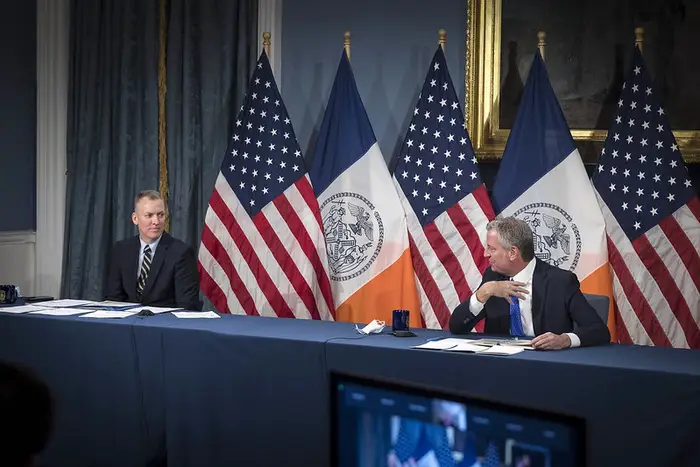 Mayor Bill de Blasio sits near a long conference table on the right as NYPD Commissioner Dermot Shea sits all the way to the left side of the conference table.